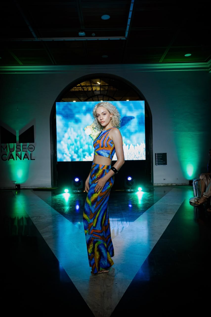 Modelo con top y pantalón de estampado azul y naranja sobre una pasarela iluminada, con una pantalla y el arco del Museo del Canal al fondo