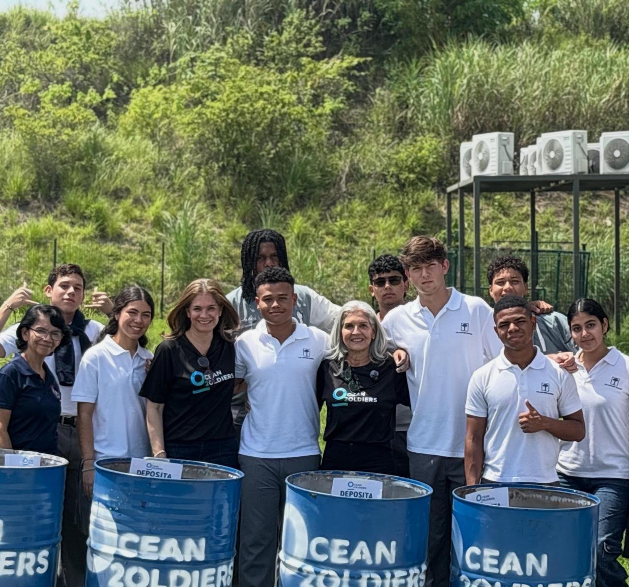 Ofelia Mercedes Méndez rodeada de un grupo de jóvenes estudiantes, detrás de botes de reciclaje azules con el logotipo de Ocean Soldiers