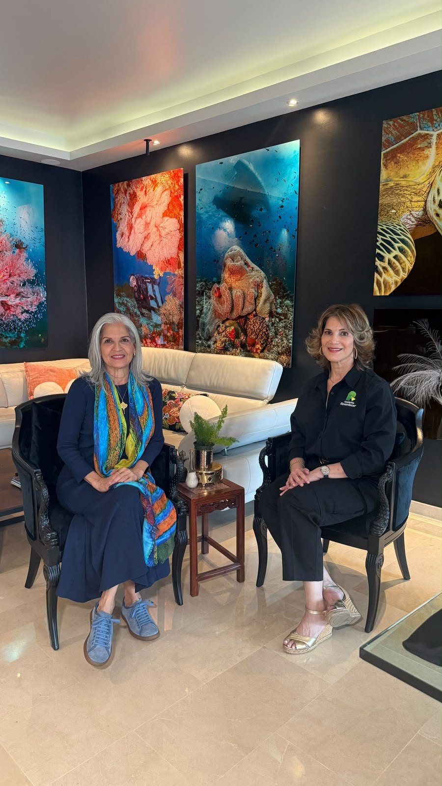 Ofelia Mercedes Méndez, con pañuelo de colores, sentada junto a Maricarmen, representante de Recimetal y ReverdecER, en una sala decorada con grandes fotografías submarinas de corales y una tortuga