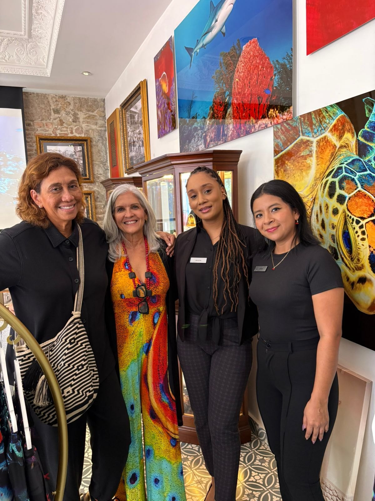 Ofelia Mercedes Méndez, con vestido largo de estampado en colores vivos, junto a tres integrantes del equipo en la sala de exhibición, rodeadas de fotografías submarinas de gran formato