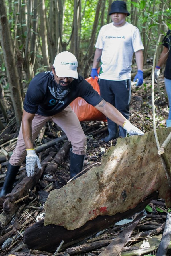 Un voluntario con gorra y guantes levanta una lámina metálica oxidada entre la vegetación, mientras otro sostiene una bolsa de recolección al fondo